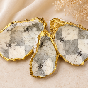 Decorative gold-rimmed ceramic shells with a black-and-white checker pattern resting on beige fabric and dried flowers in the corner.