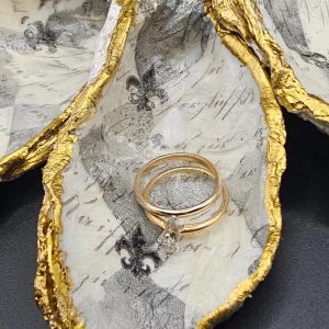Open egg-shaped jewelry box with a distressed gold rim, lined with patterned fabric, holding three gold rings including a diamond pendant ring