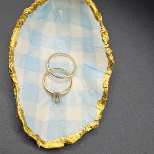 Oval container lined with blue-and-white checkered fabric and a crumpled gold edge, holding two silver rings with a small pendant between them. (Informative: shows jewelry display object.)